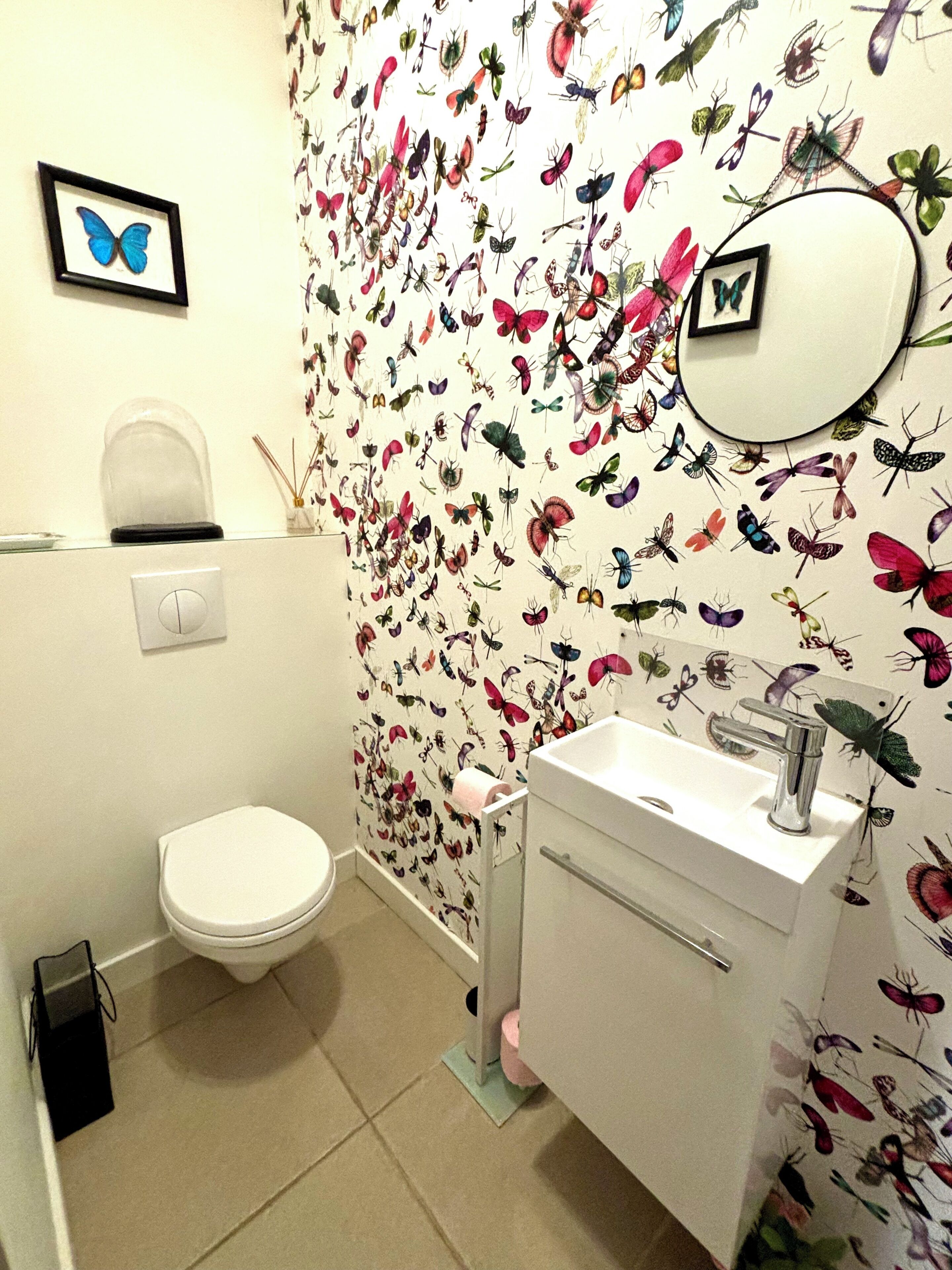 Une salle de bain comprenant des toilettes, un lavabo et un miroir. Les murs sont décorés d’un papier peint représentant des papillons et des insectes.