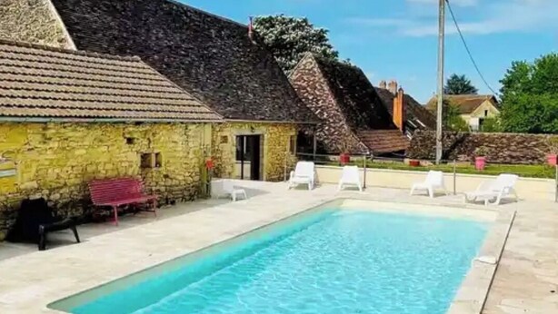 Una piscina con sillas reclinables blancas, un edificio de piedra con tejado de baldosas y un cielo azul despejado.