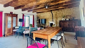 Un interior rústico con vigas de madera a la vista, una zona de comedor con mesa y sillas de madera, un muro de piedra y un televisor.