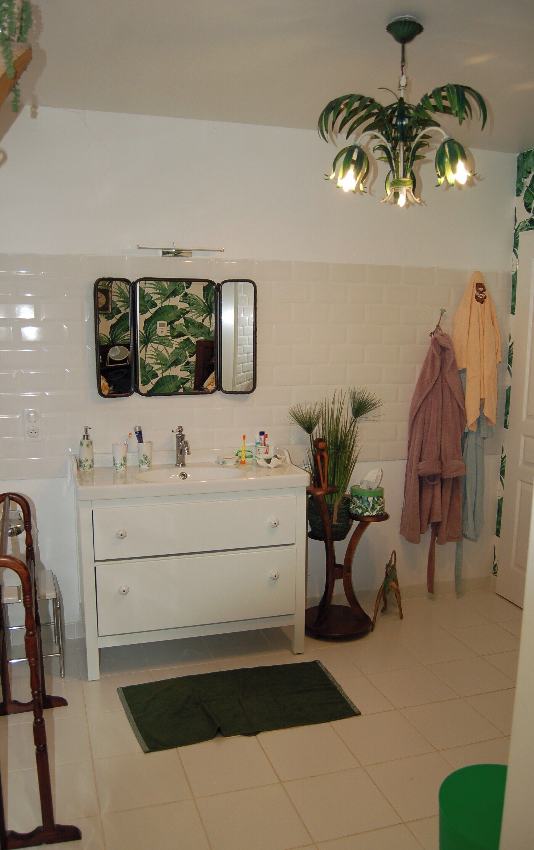 Une salle de bain avec un meuble-lavabo blanc, un miroir, un lavabo avec des articles de toilette, une plante verte, une chaise en bois et un peignoir accroché au mur.