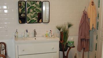 Une salle de bain avec un meuble-lavabo blanc, un miroir, un lavabo avec des articles de toilette, une plante verte, une chaise en bois et un peignoir accroché au mur.