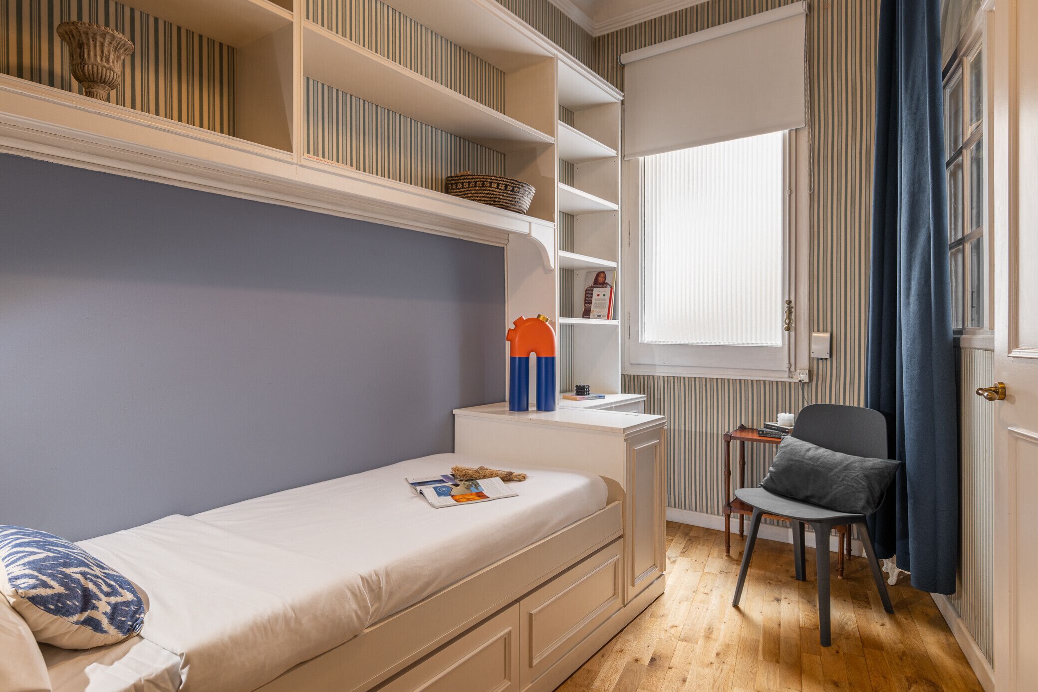 A compact bedroom with a single bed, a small desk, and a chair. There is a window with curtains, a radiator, and a shelf with decorative items.