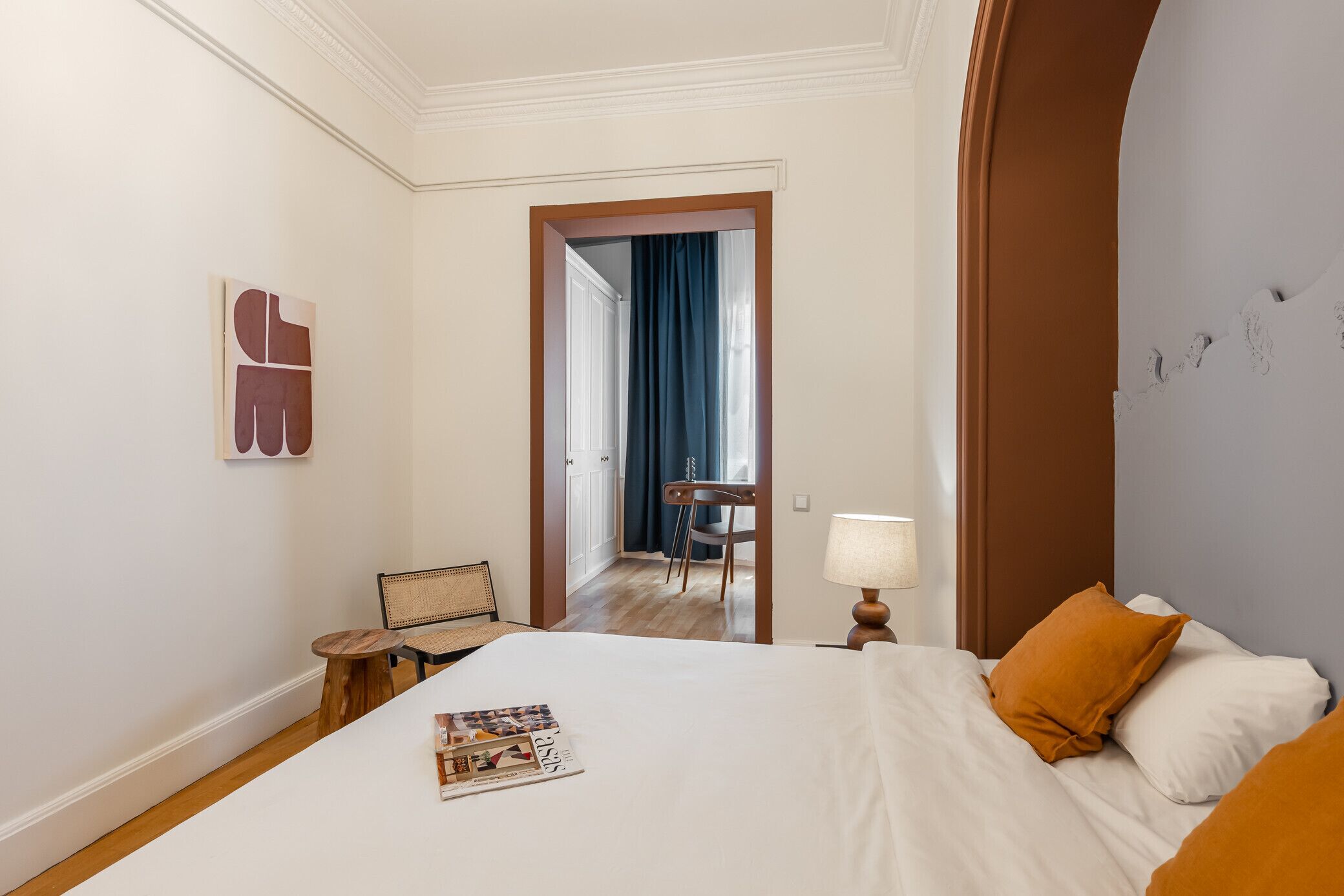 A hotel room with a bed, a small table with a lamp, a chair, and a door leading to another room.