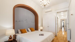 Une chambre à coucher avec un grand lit, une table de chevet en bois, un lustre et un couloir menant à d’autres pièces.