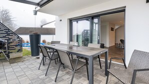 En uteplats med ett bord och stolar, en grill och en stor skjutdörr i glas som leder in till ett inomhusutrymme.