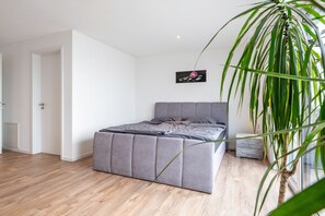 Une chambre à coucher avec un lit recouvert d’un tissu gris, une petite table de chevet, une commode, une plante en pot et un tableau accroché au mur.