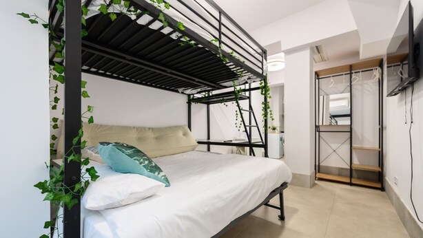 A bunk bed with white bedding, a beige pillow, and a teal pillow. There is a small table beside the bed with a plant on it. The room has white walls, a wooden door, and a TV mounted on the wall. - (RJS3) Cozy Studio in Salamanca District (Madrid)
