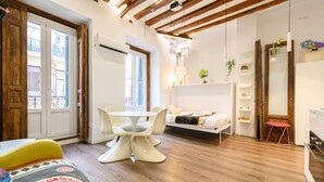 Habitación pequeña con cama, una mesita y una silla. Se aprecia una ventana con vistas a edificios.