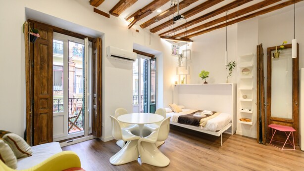 Un espacio habitable compacto con techo de madera, una cama blanca, una mesita redonda y un balcón con vistas.