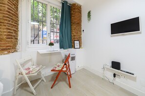 Ein Zimmer mit Fenster, weißem Schreibtisch, Stuhl, Fernseher und Heizung.