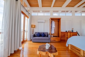 Un'ampia stanza con soffitto in legno, un divano grigio con cuscini blu, una cassettiera in legno e un letto con copripiumino blu.