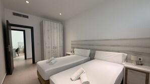 Ein Hotelzimmer mit zwei Betten, einem Kleiderschrank, einem Nachttisch mit Lampe und Blick durch eine offene Tür.