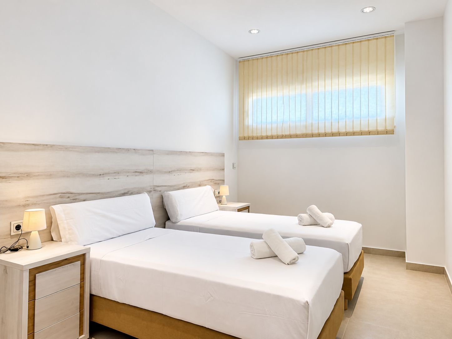 Una habitación de hotel con dos camas, una mesita de noche con lámpara y una ventana con persianas.