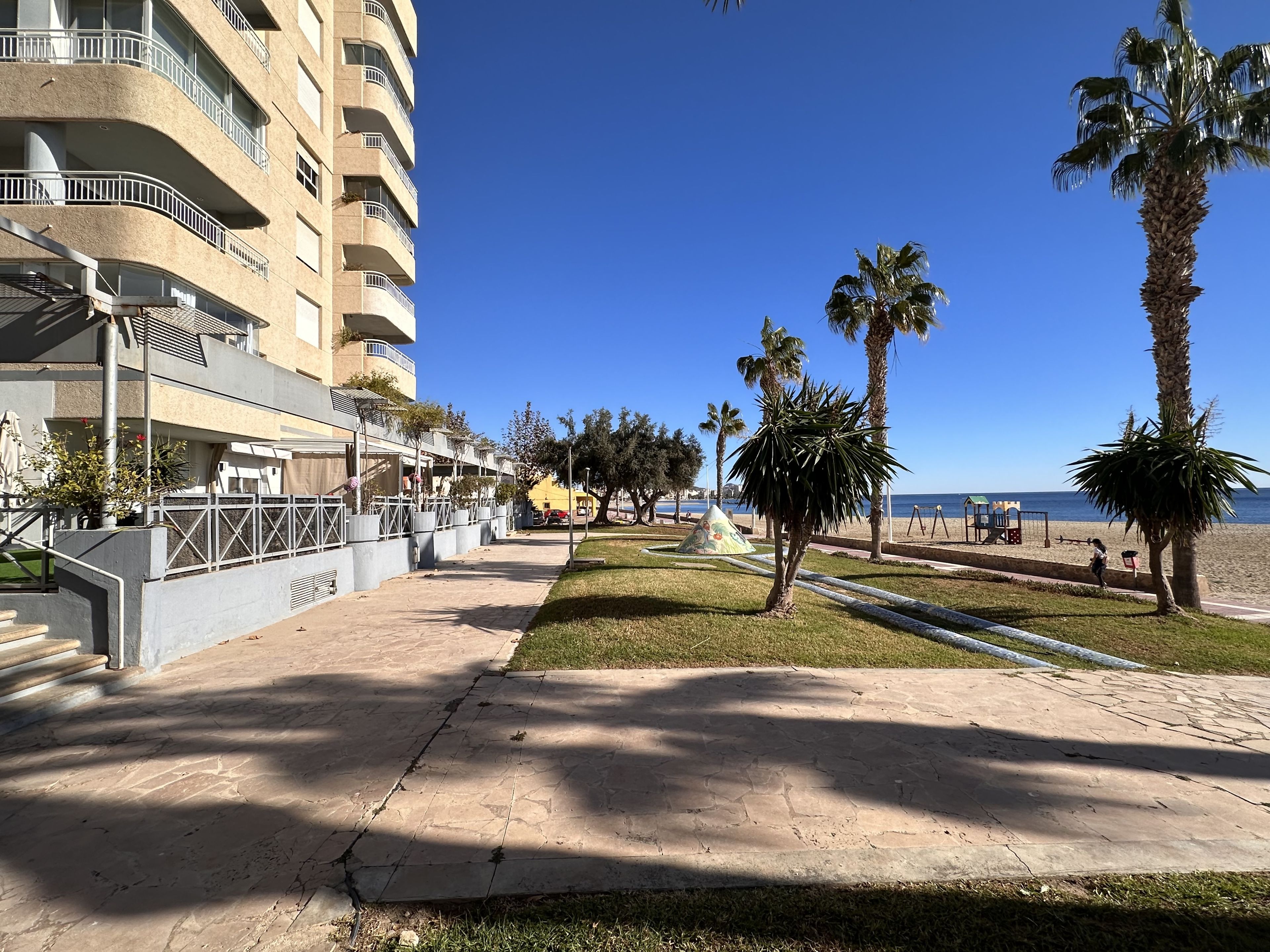 Un paseo marítimo frente a la playa con un edificio de varios pisos, palmeras y un cielo despejado.