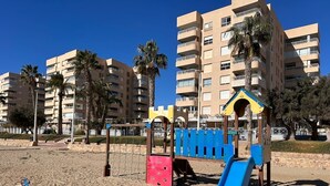A playground with a slide and swings on a sandy beach, surrounded by palm trees and residential buildings. - Apartment 65B · Oceanfront with Private Terrace (Aguilas)