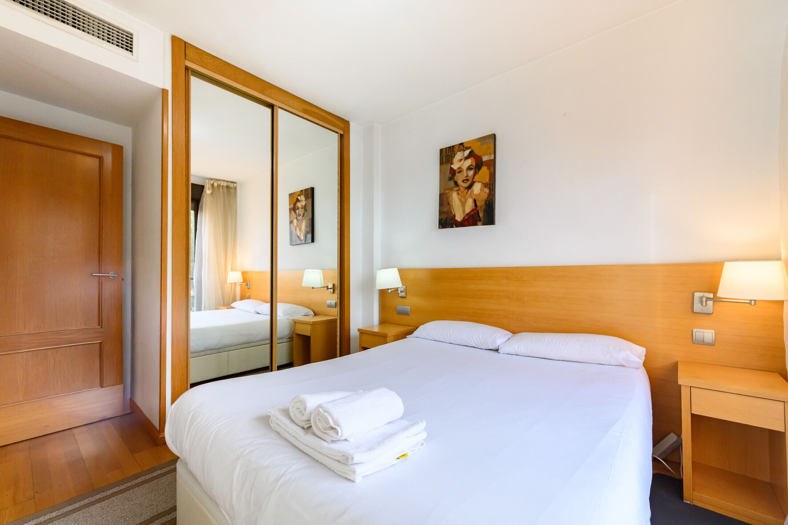A hotel room with a large bed, a wooden wardrobe, a bedside table with a lamp, and a painting on the wall.