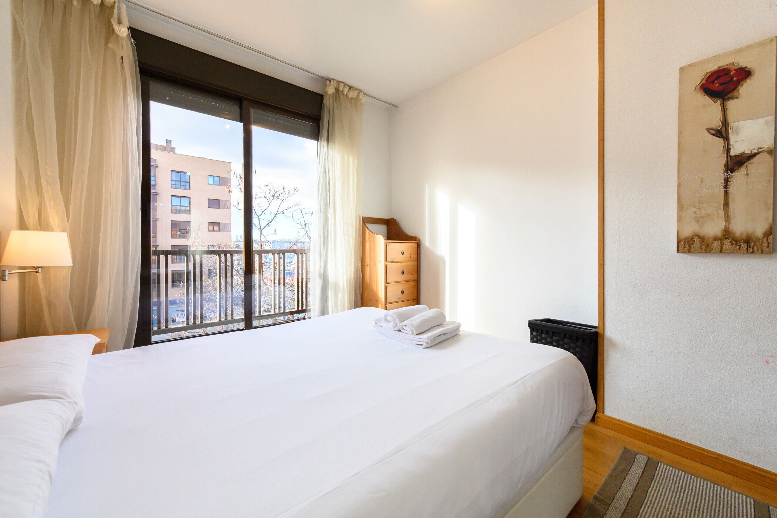 Habitación de hotel con una cama grande, un aparador de madera, una ventana con cortinas, un cuadro de una rosa y vistas a edificios.