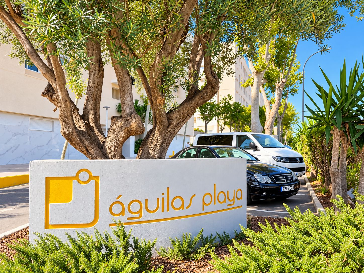 Señalización para 'Águilas playa' con un área de estacionamiento y una calle arbolada.