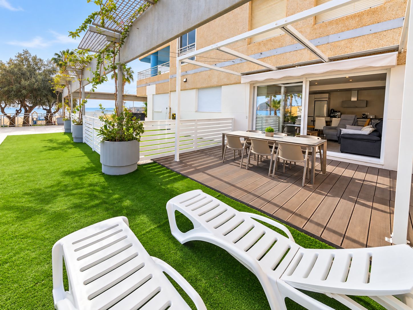A modern outdoor patio with a wooden deck, white lounge chairs, a dining table, and potted plants.