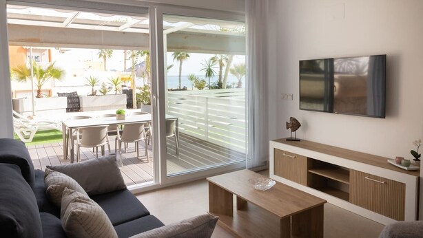 Un salon moderne avec un canapé, une table basse et un téléviseur. Une porte-fenêtre coulissante donne accès à un balcon aménagé avec des meubles d’extérieur et des plantes.