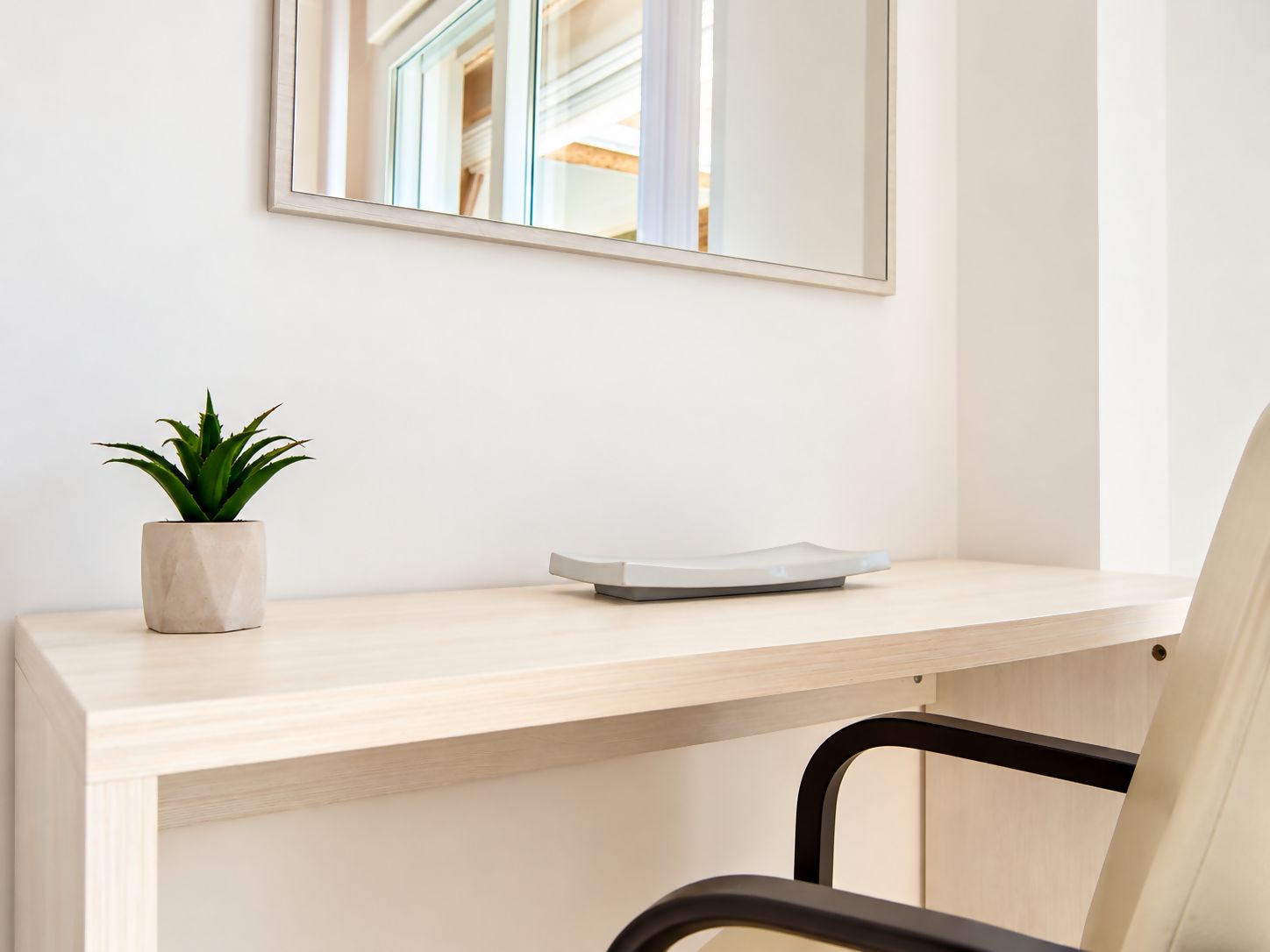 A minimalist desk with a potted plant and a white object, possibly a speaker, against a plain wall with a mirror reflecting a room.