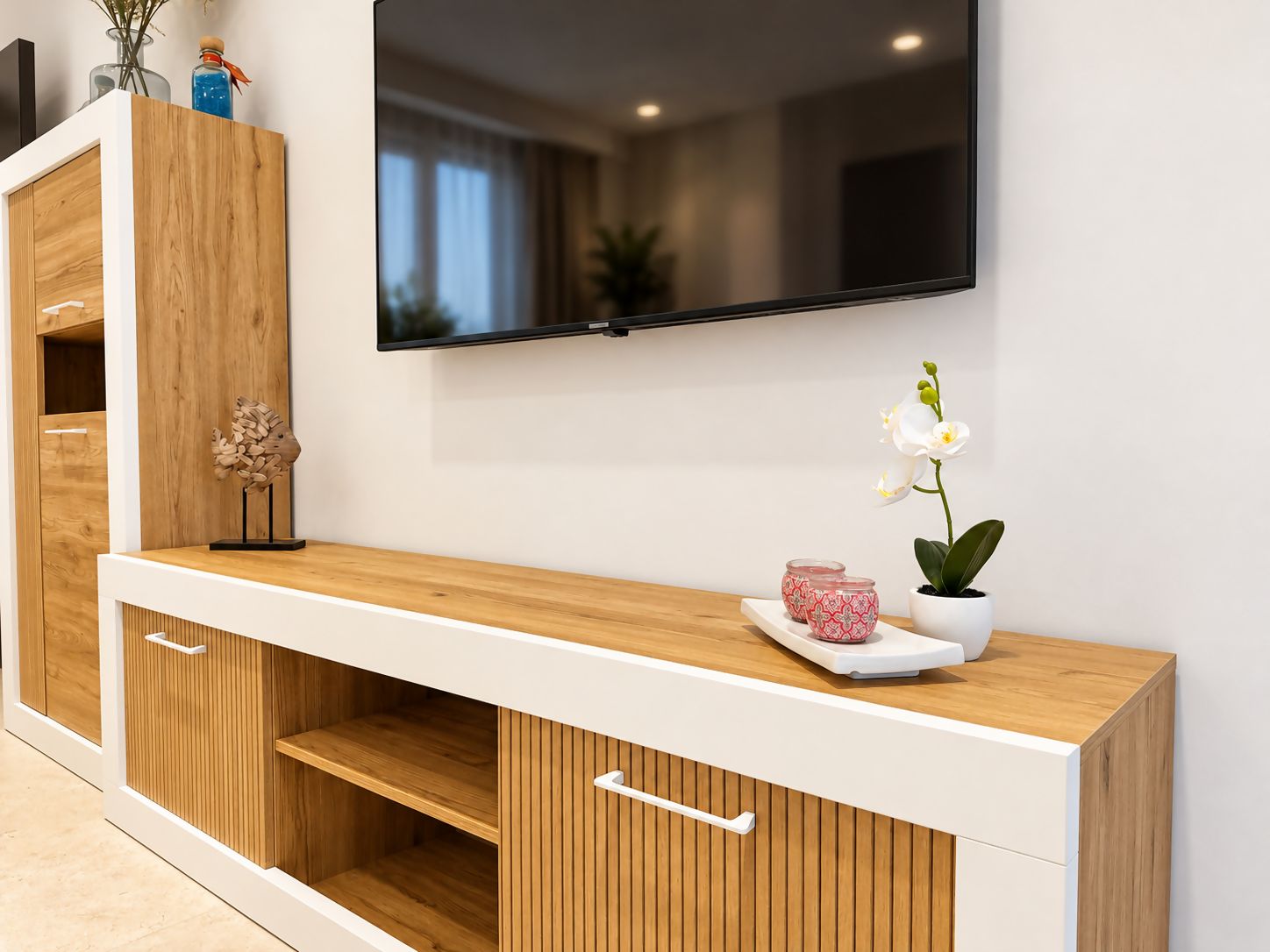 Un salon moderne avec un téléviseur à écran plat fixé au mur, des meubles en bois dotés d’étagères ouvertes et une orchidée en pot posée sur la console.