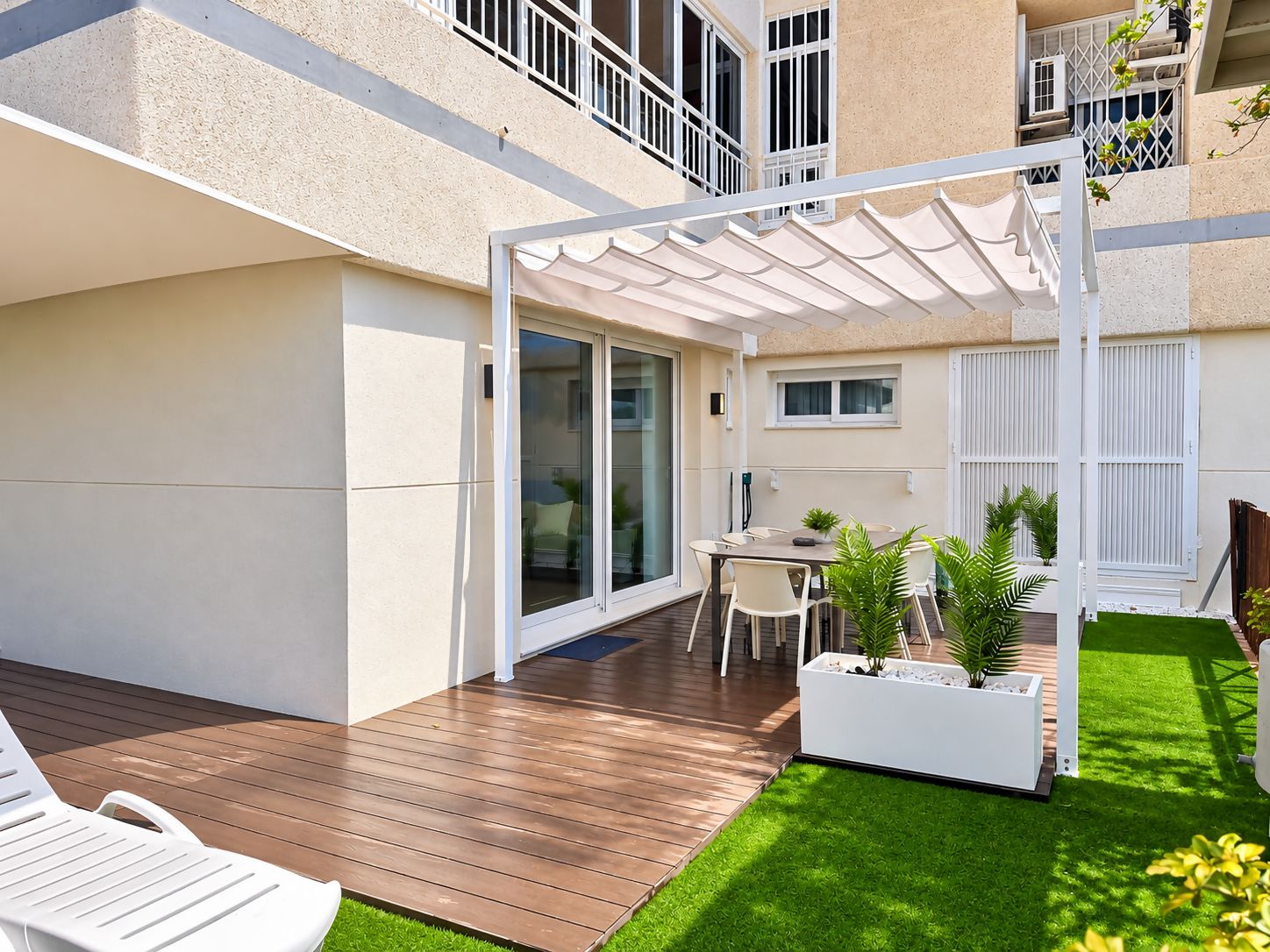 A modern outdoor patio with a wooden deck, a covered seating area, and a small garden with potted plants.