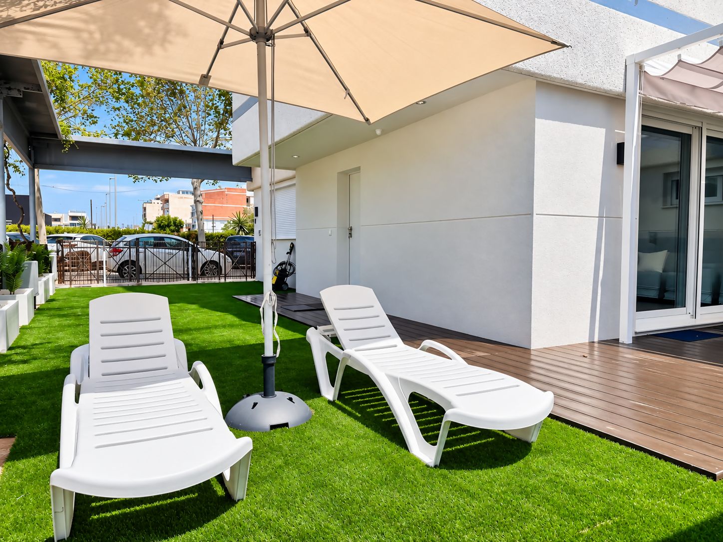Eine Terrasse mit zwei weißen Liegestühlen, einem Tisch und einem beigefarbenen Sonnenschirm.