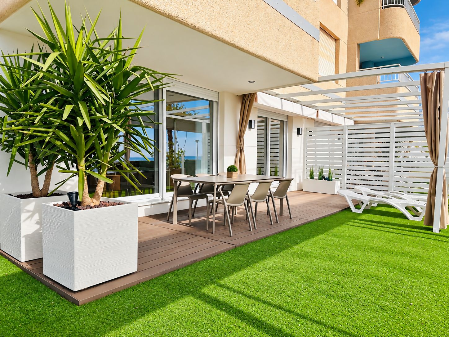 Een modern terras buiten met een eettafel en stoelen, omgeven door een witte hek en een grote plant.