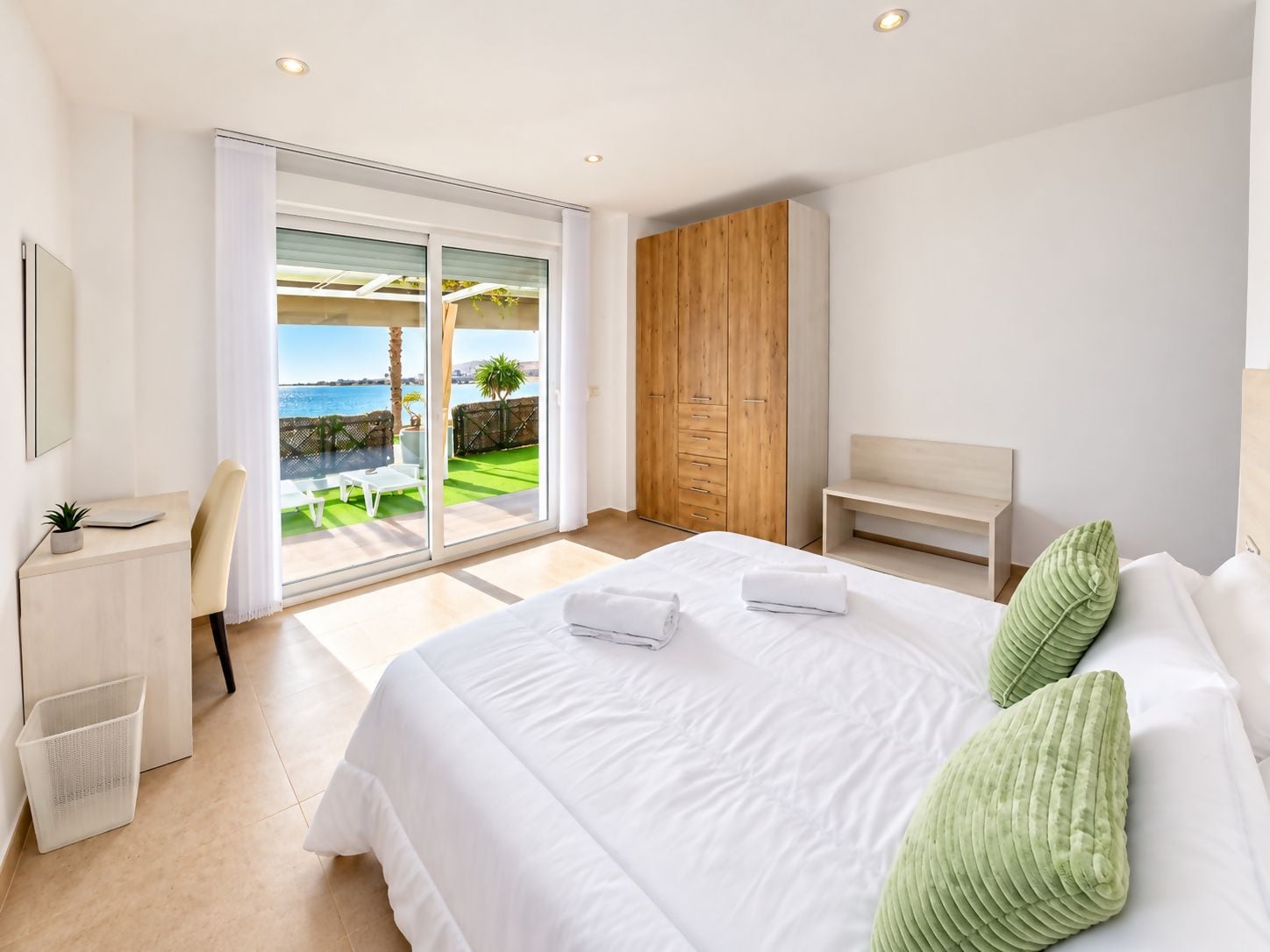 A modern bedroom with a large bed, a wooden wardrobe, a desk with a chair, and a view of the sea through a sliding door.