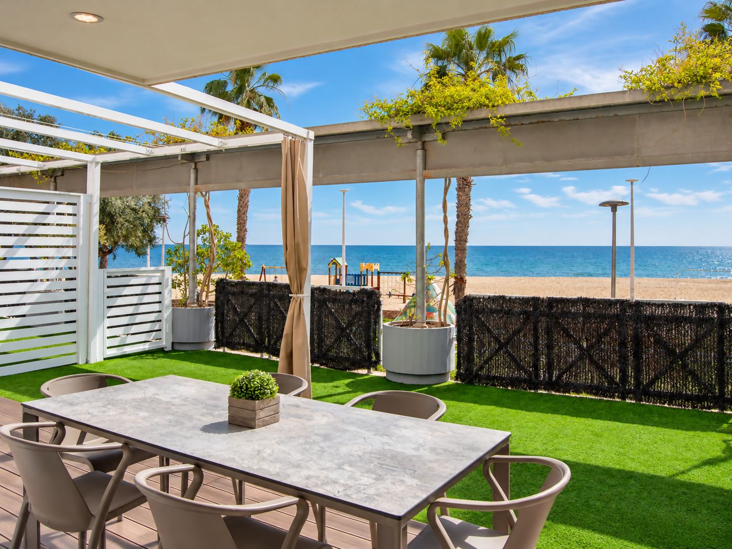 An outdoor patio with a table and chairs, a view of the beach, and a pergola with plants.