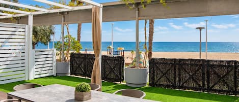 An outdoor patio with a table and chairs, a view of the beach, and a pergola with plants.