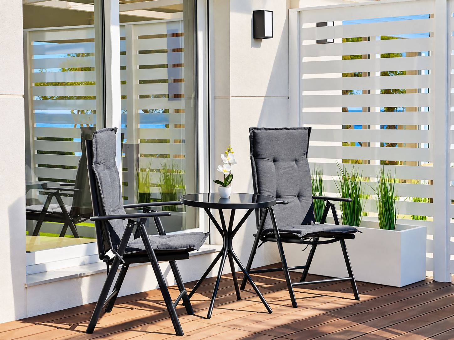 Ein Sitzbereich im Freien mit zwei Stühlen und einem Tisch, ein weißer Pflanzkasten mit Pflanzen und ein weißer Zaun mit horizontalen Lamellen.