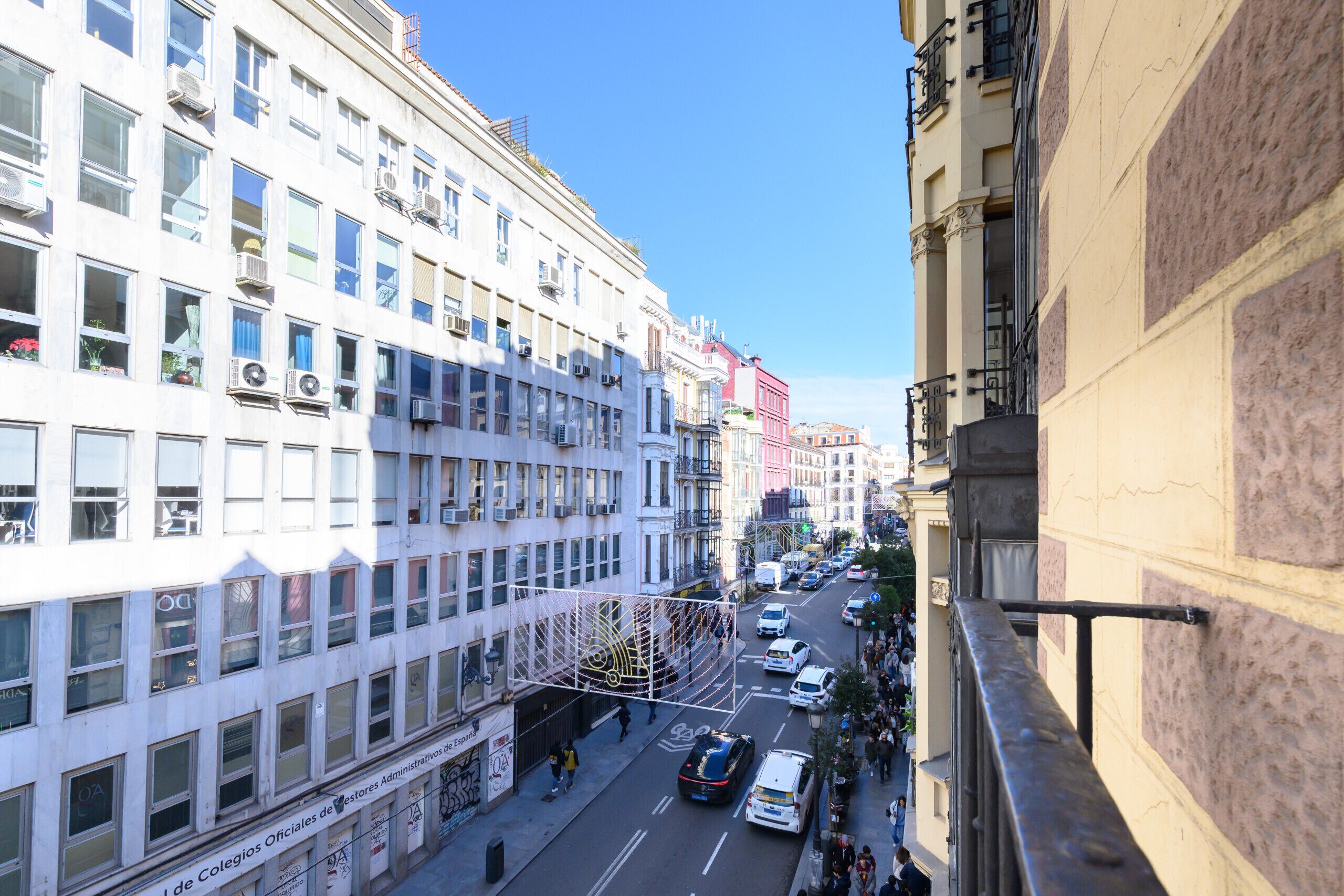 Un edificio de varios pisos con una fila de ventanas en la planta baja, un paseo peatonal y una calle con coches y gente.