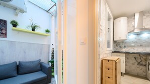 Un espace de vie compact comprenant un canapé, un petit meuble en bois, un lave-linge et un coin cuisine avec un dosseret carrelé.