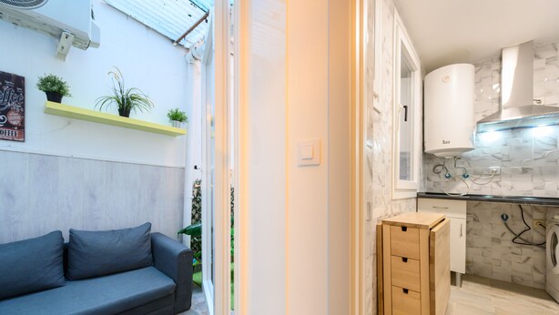 Ett kompakt bostadsutrymme med en soffa, ett litet träskåp, en tvättmaskin och en köksdel med kakel på väggen bakom arbetsbänken.