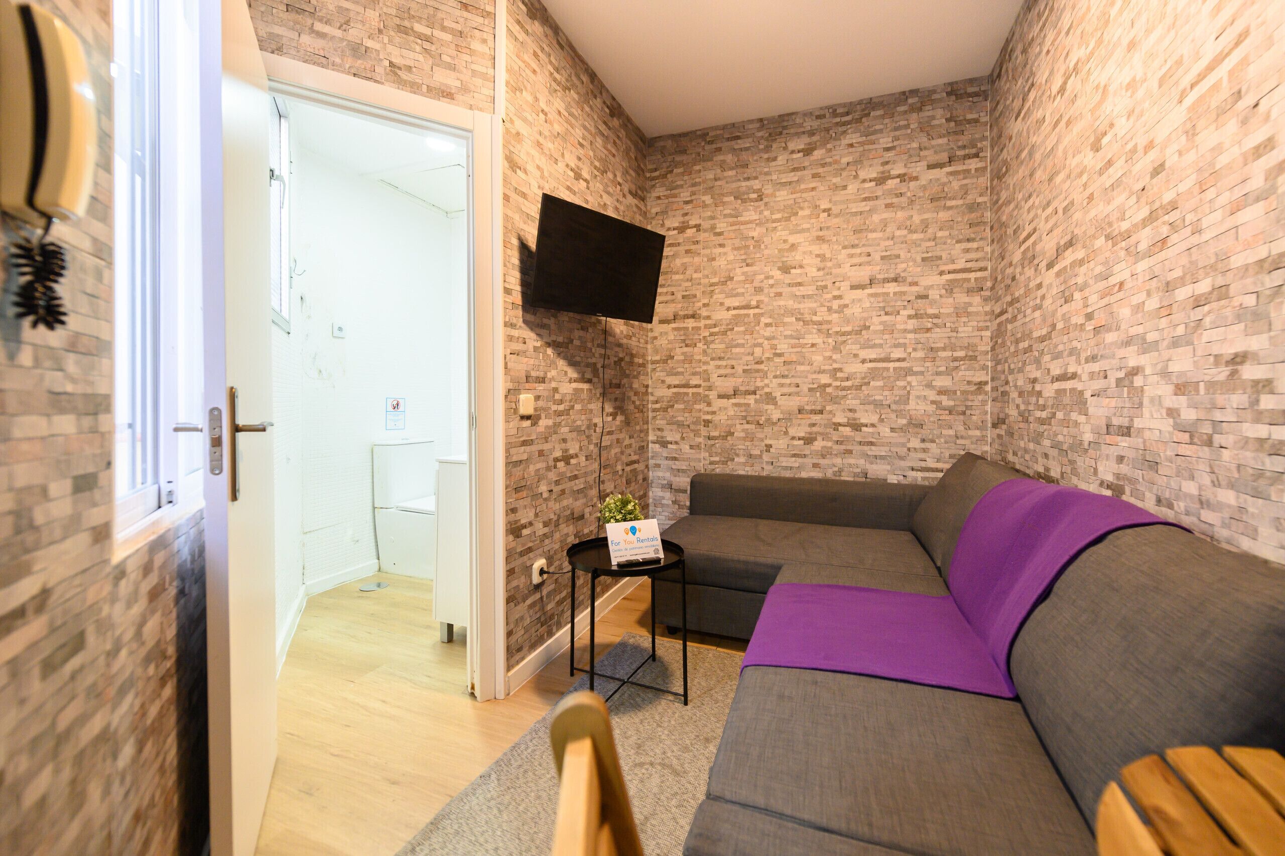 A room with a brick-patterned wall, a gray sofa with a purple throw, a mounted TV, and a small table with a sign.