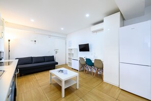 Ein modernes, helles Wohnzimmer mit einer Couch, einem Couchtisch, einem Fernseher und einer Küchenzeile mit Kühlschrank und Schränken.