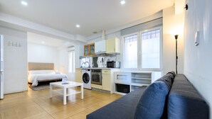 Een moderne, goed verlichte kamer met een bed, een slaapbank, een klein tafeltje en een kitchenette met een magnetron en oven.