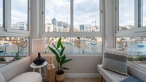 Een kamer met een groot raam dat uitkijkt op een jachthaven en gebouwen, een bank met kussens, een lamp en een potplant.