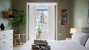 Een slaapkamer met een bed, een kaptafel, een kamerplant, een nachtkastje en een uitzicht op gebouwen door een geopende deur.