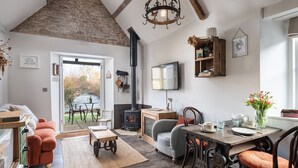 A cozy living room with a stone ceiling, a fireplace, a television, and a dining area with a table and chairs. - Filly Island - sleeps 2 guests  in 1 bedroom (South Cerney)