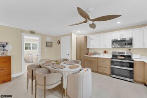 A modern kitchen with white cabinets, a stainless steel microwave, and an oven. There is a dining area with a table and chairs, and a ceiling fan above. - Gulf Westwind 3-1. Across the street from Beach! Resort style pool. (Fort Myers Beach)