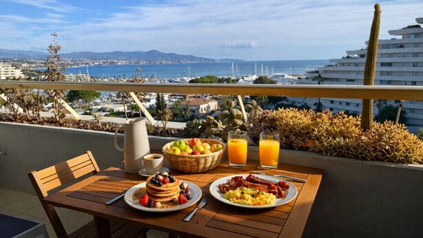 Ein Balkon mit Frühstücksangeboten, darunter Pfannkuchen, Eier und Saft, mit Blick auf eine Küstenstadt.