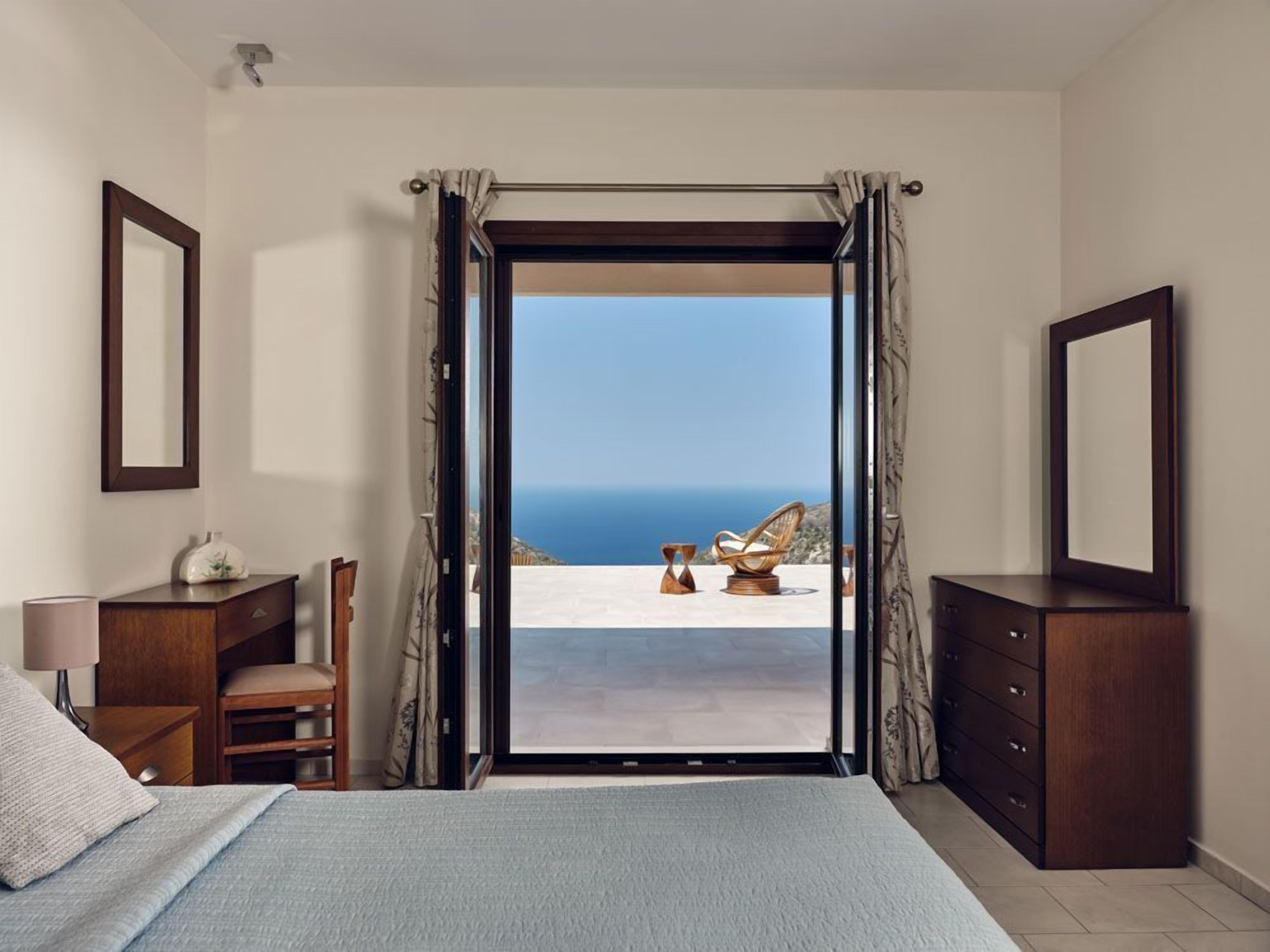 A bedroom with a large bed, a dresser, a chair, and a view of the sea.