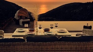 Mesa de jantar posta com pratos, copos e um vaso de flores, com vista para o pôr do sol sobre o mar.