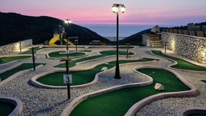 Um campo de mini golfe ao entardecer, com vista panorâmica do mar e das colinas ao fundo.