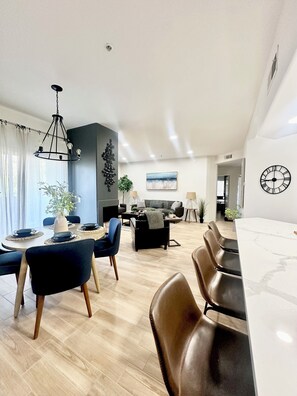 En moderne stue med spiseplads, med en lysekrone, sofasæt og et stort ur på væggen.