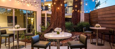 An outdoor patio with round tables and chairs, string lights, and large trees.