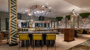 Een moderne bar met een centraal gelegen toog, diverse zitplaatsen waaronder barstoelen en hoge kinderstoelen, en een decoratieve plafondconstructie.